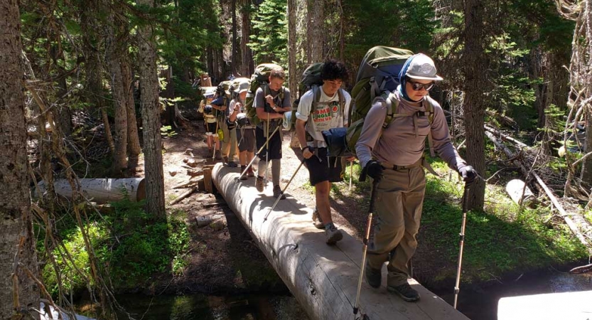 A group of teens wearing backpacks and using trekking poles hike over a wood plank